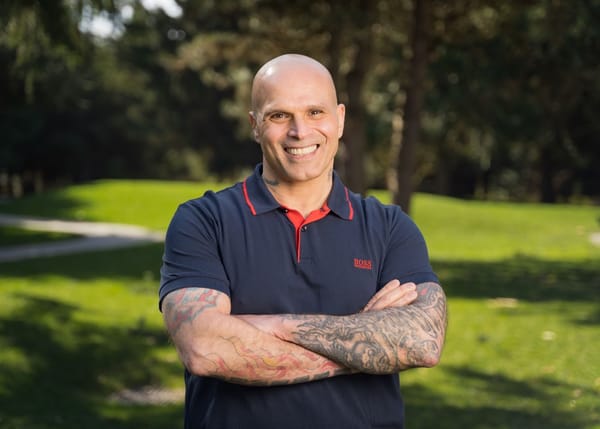 A man with a big grin stands with tattooed arms crossed with a green grassy meadow behind.
